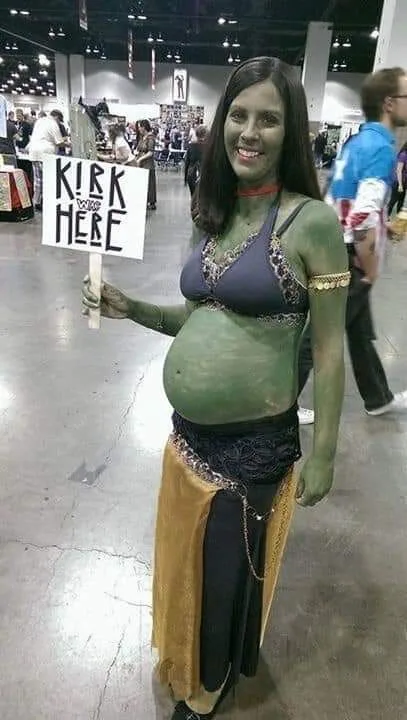 A woman dressed as a green-skinned alien with a visibly pregnant belly holds a sign that reads 'KICK YOURS HERE'. She wears…