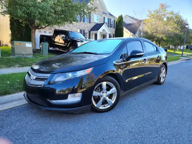 A shiny black Chevrolet Volt is parked on a residential street, with a large black pickup truck visible in the background…