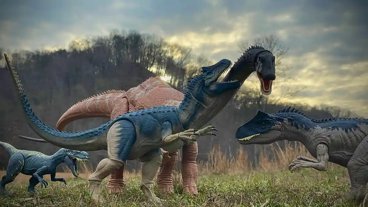 Four plastic dinosaur toys are arranged in a grassy field with a wooded hill in the background, appearing to be in a…