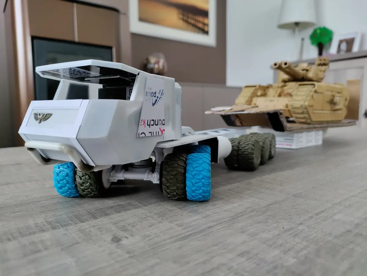 A white toy truck with blue and green tires is pulling a tan toy tank on a wooden floor. The truck has a sticker that reads…