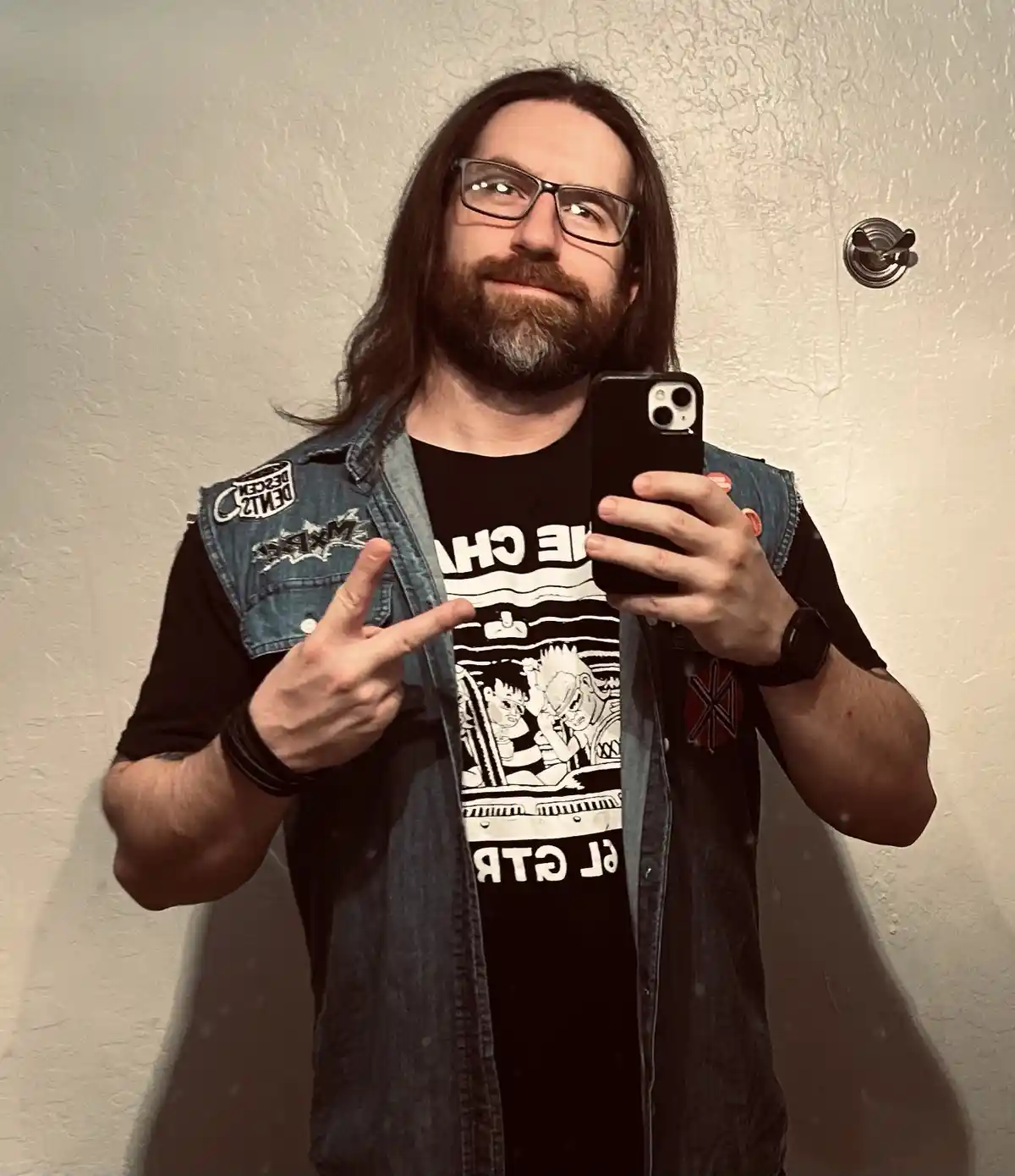 A man with long hair, glasses, and a beard takes a mirror selfie holding a black smartphone. He wears a denim vest over a…