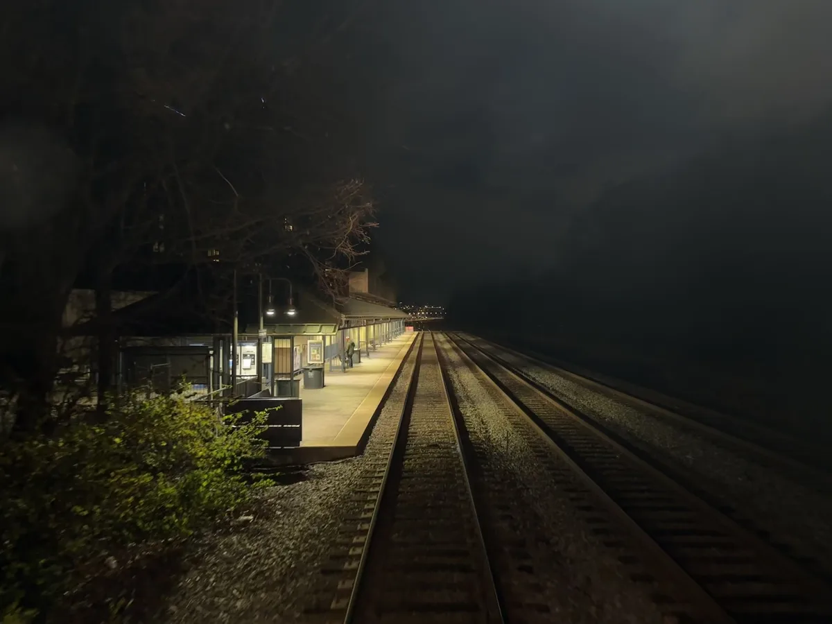 A nighttime photo of an empty train station platform with tracks receding into the dark, illuminated by station lights. A…