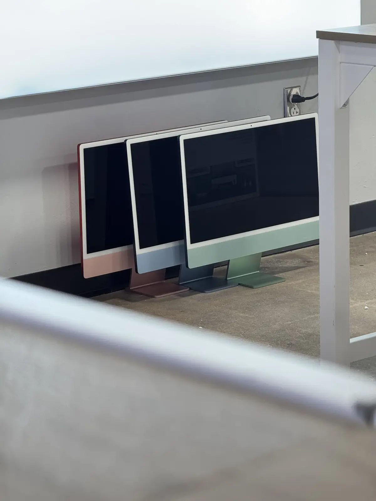 Three Apple iMac computers in pink, blue, and green are lined up with their screens off, viewed from behind a white barrier…