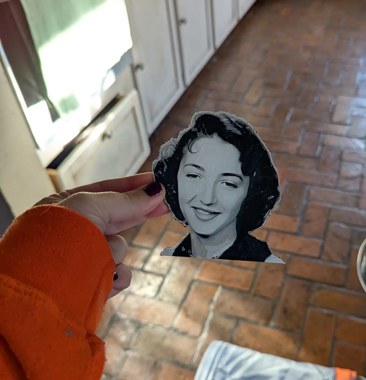 A hand with dark red nail polish holds up a crudely cut-out, black-and-white portrait of a smiling woman, likely the…