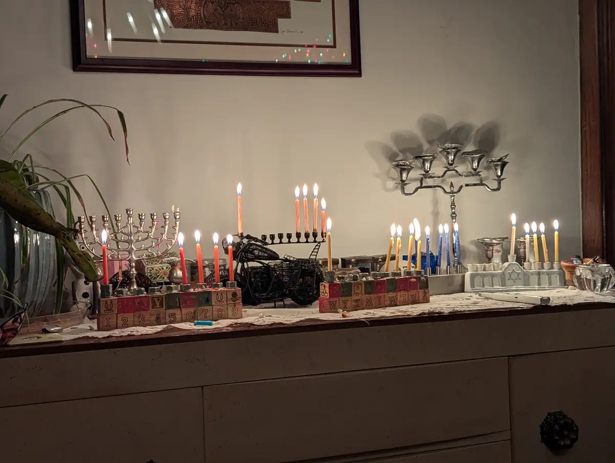 A cluttered dresser top displays multiple lit menorahs of varying styles and sizes, including a decorative one made from…
