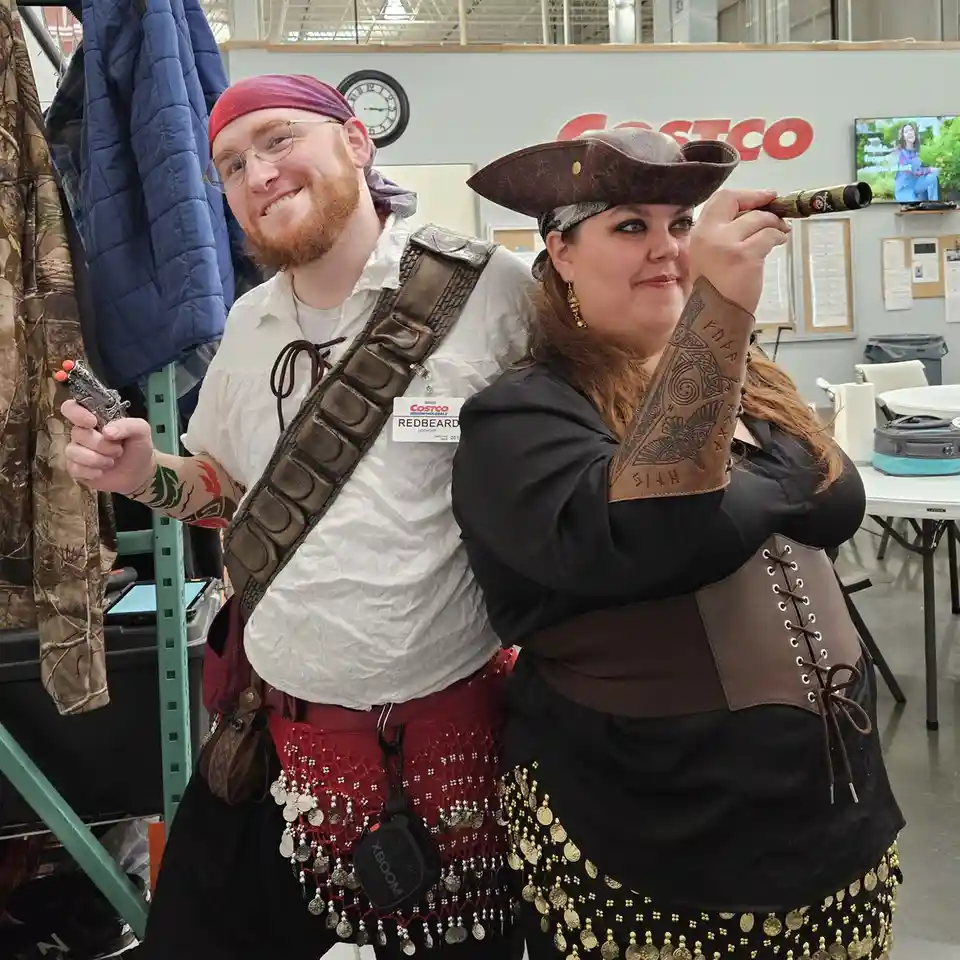 Two people dressed as pirates pose inside a Costco warehouse, with the man wearing a red bandana and a name tag that reads…