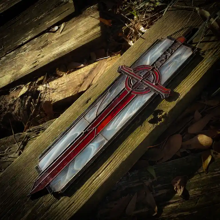 A stained-glass sword pendant with red and clear glass pieces, outlined in black lead, rests diagonally on weathered wooden…