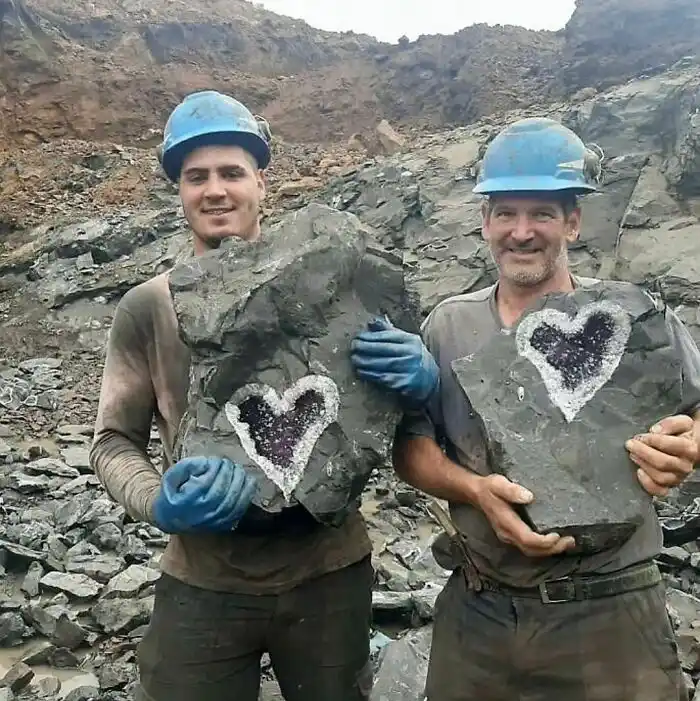 Two men in blue hard hats and work clothes stand in a rocky quarry, each holding a large grey rock with a heart-shaped geode…