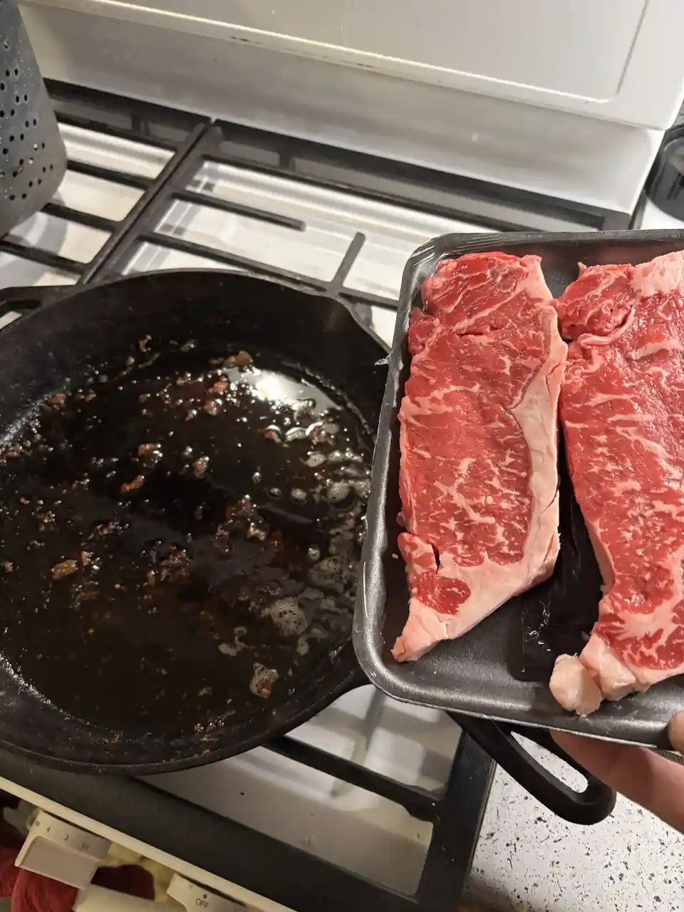 A hand holds a black plastic tray with two raw, marbled steaks over a stovetop. Beside it, a black cast-iron skillet sits on…