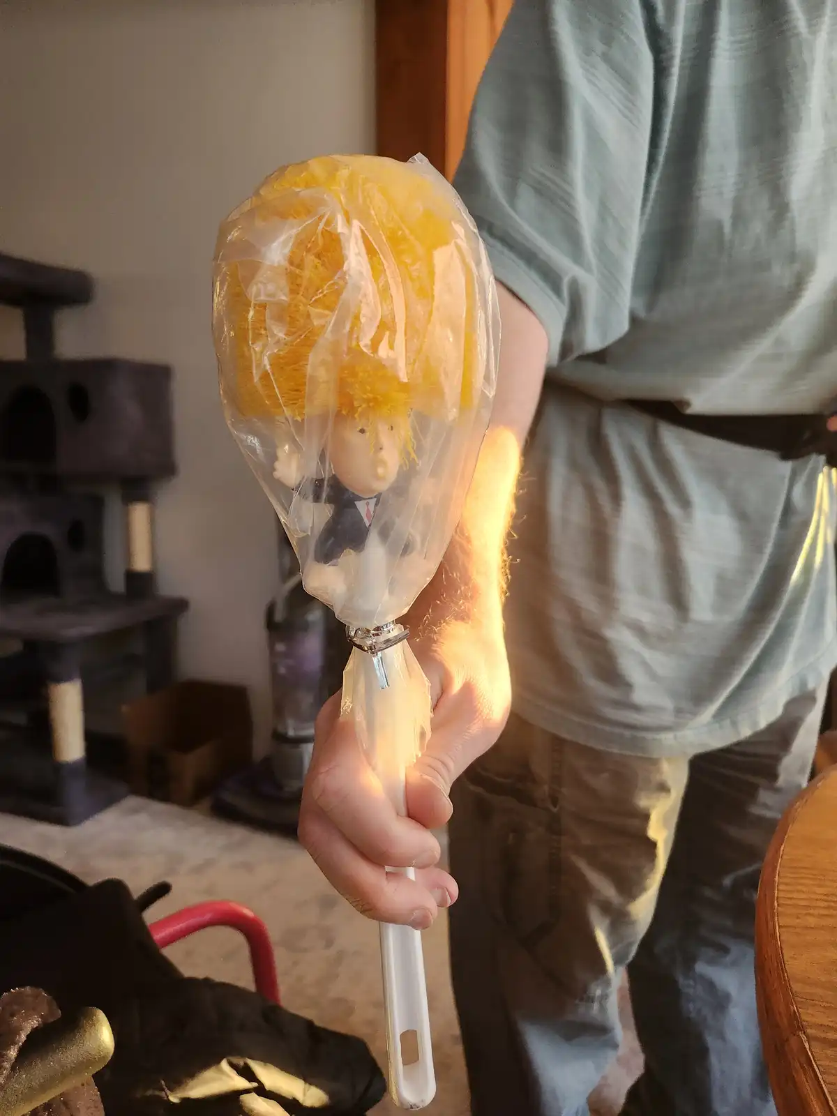 A person holds a toilet brush with a small figurine of a man in a suit and tie, wrapped in clear plastic like a lollipop…