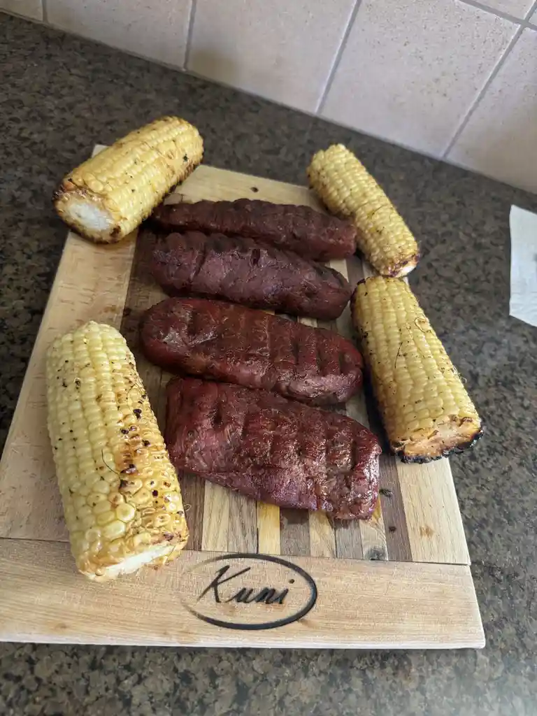 A wooden cutting board branded with 'Kuni' holds four grilled sausages and four grilled corn on the cob, arranged on a…