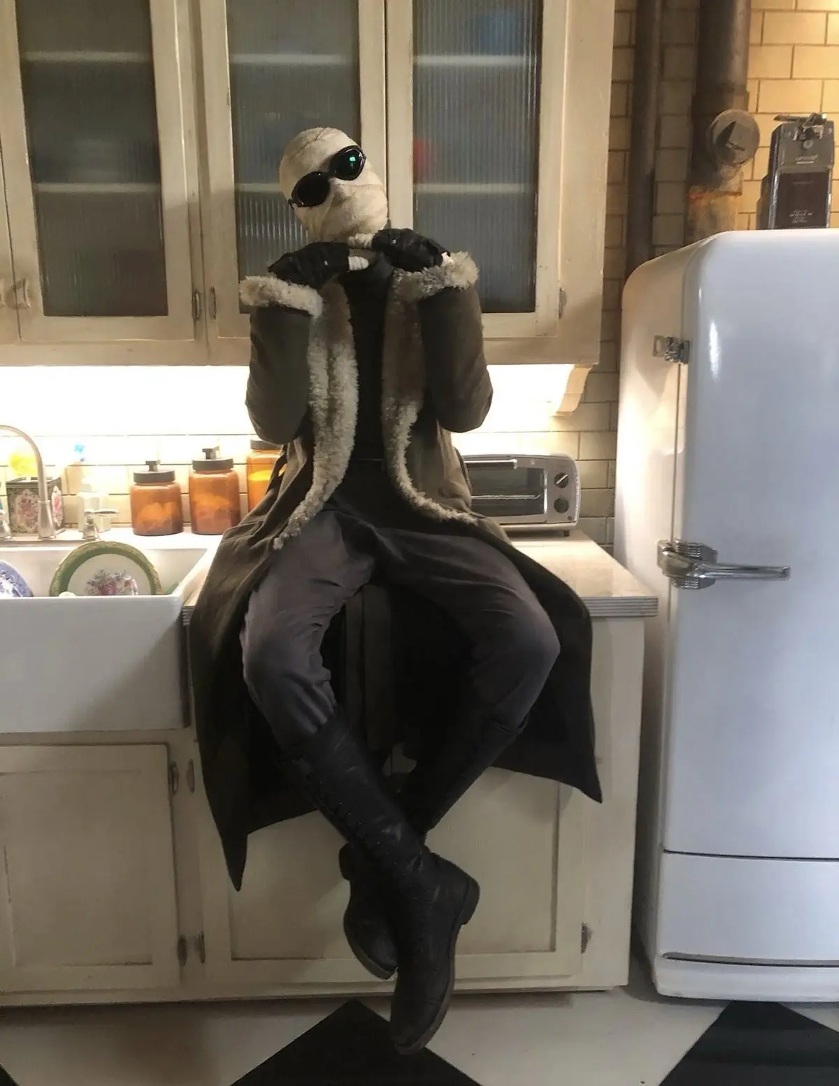 A person dressed as a mummy with a bandaged head and sunglasses sits casually on a kitchen counter, wearing a long coat with…