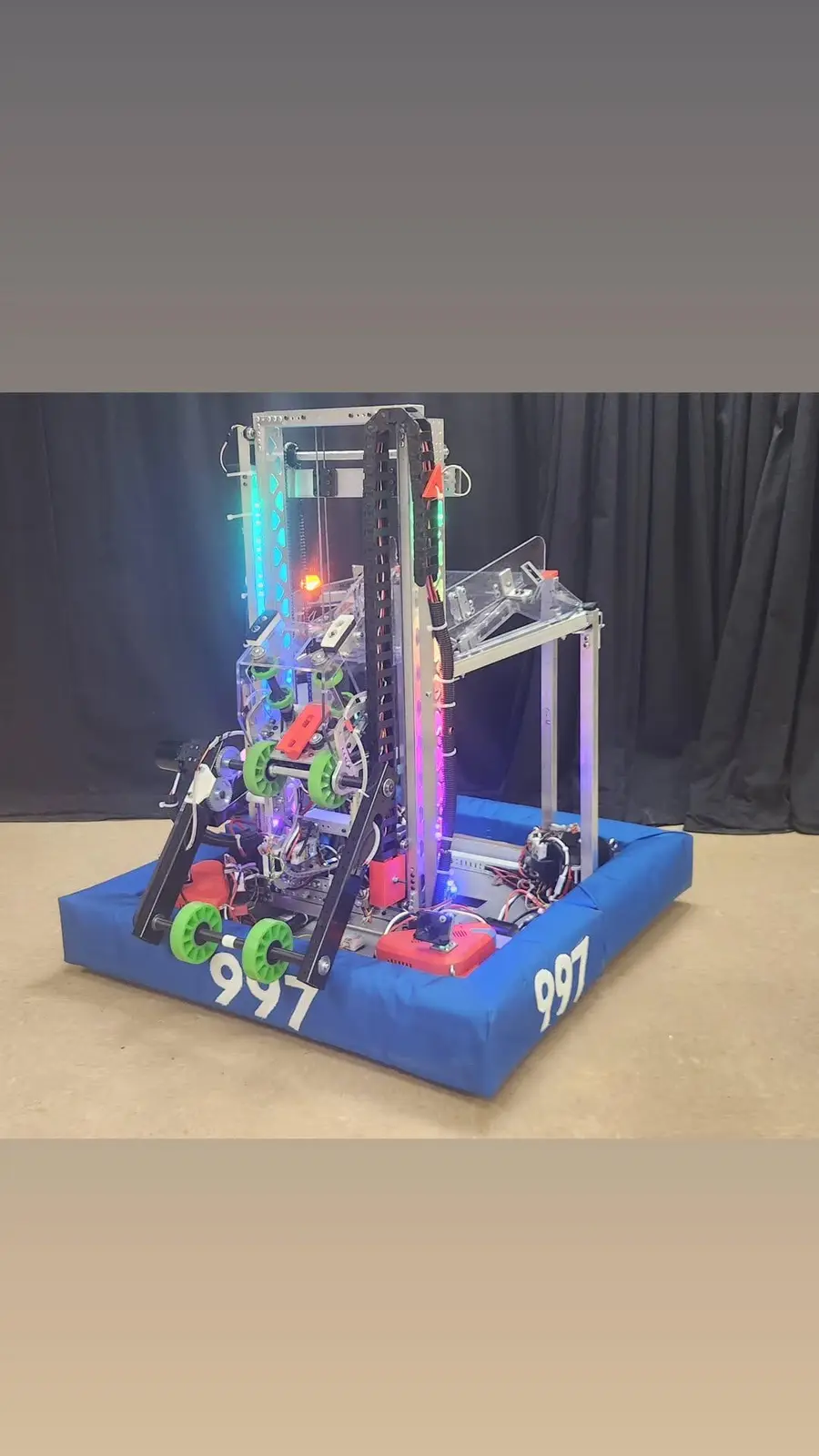 A complex, custom-built robot with a blue padded base marked with the number 997, featuring exposed wiring, green wheels…