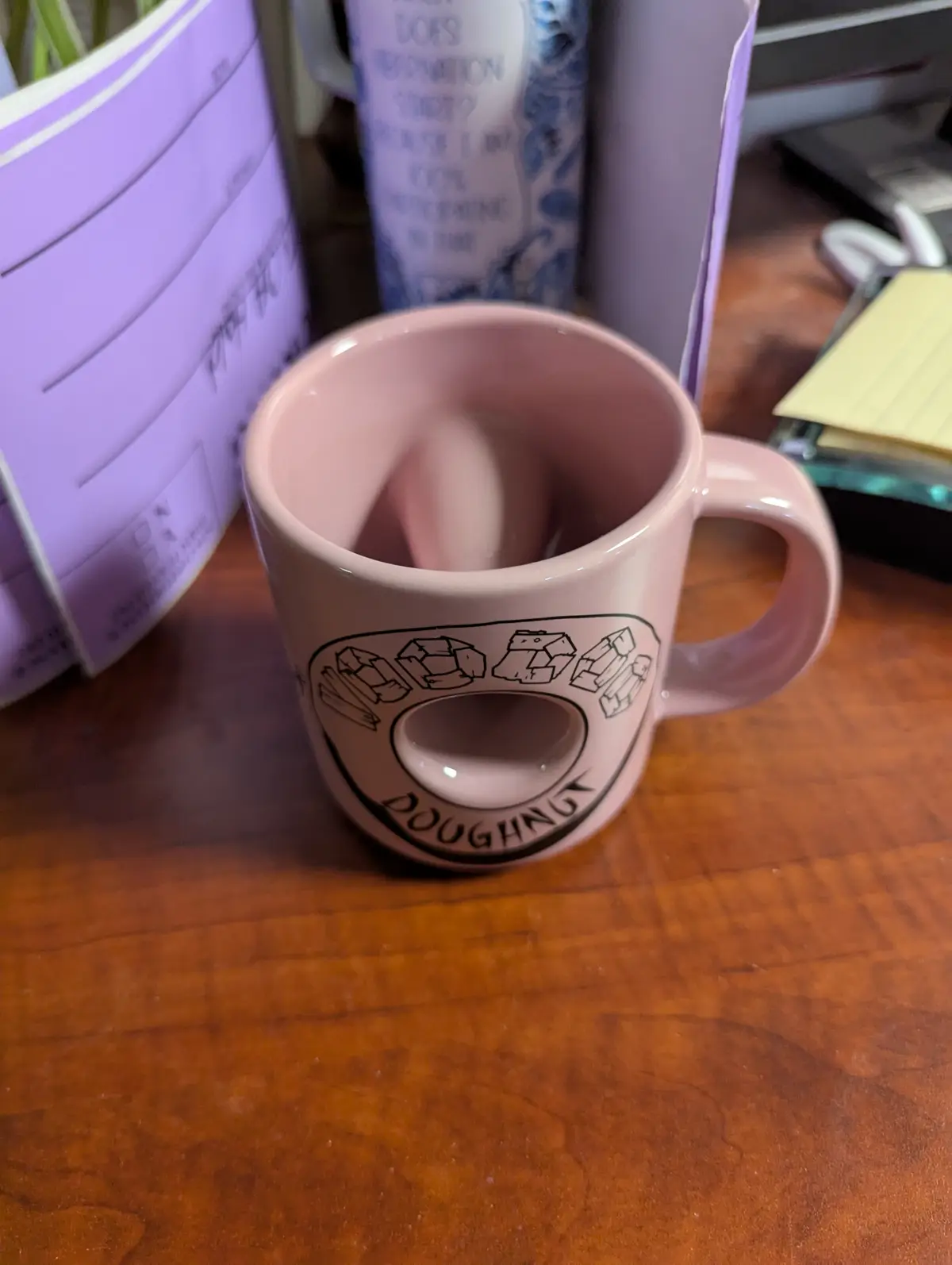 A pink ceramic mug with a handle sits on a wooden desk, featuring a black outline logo that reads 'DOUGHNUT' around a…
