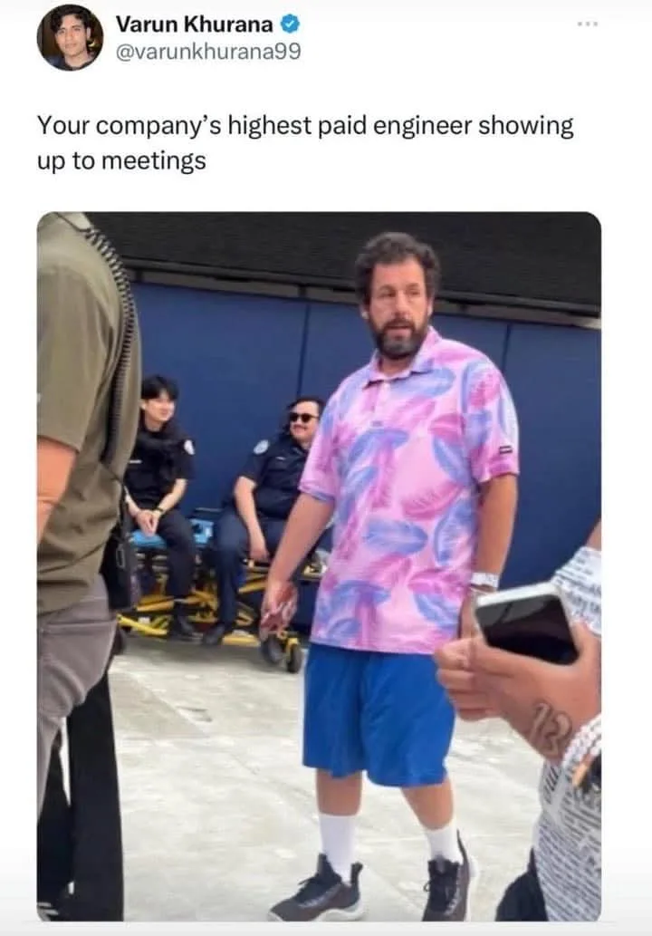 A man with a beard wearing a pink and blue patterned polo shirt and blue shorts stands in a group, looking slightly awkward…