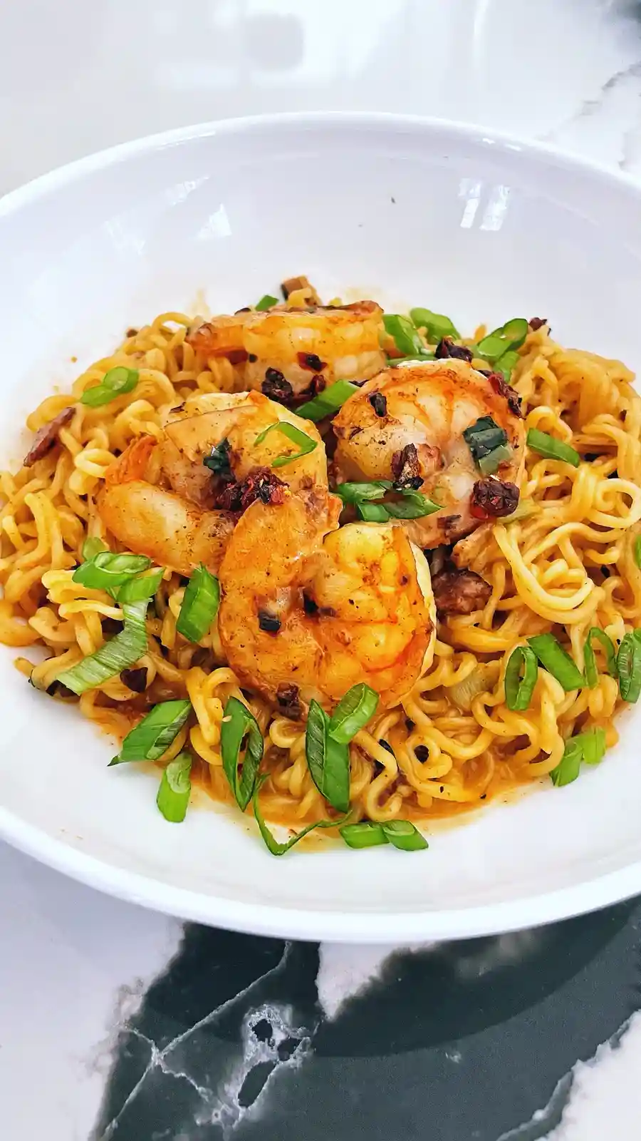 A close-up photo of a white bowl filled with yellow ramen noodles topped with several large cooked shrimp, chopped green…