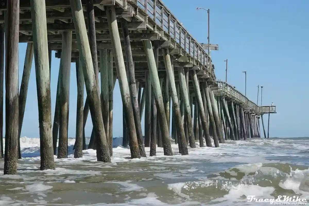 A low-angle photo shows the wooden pilings of a pier extending into the ocean under a clear blue sky, with waves crashing at…