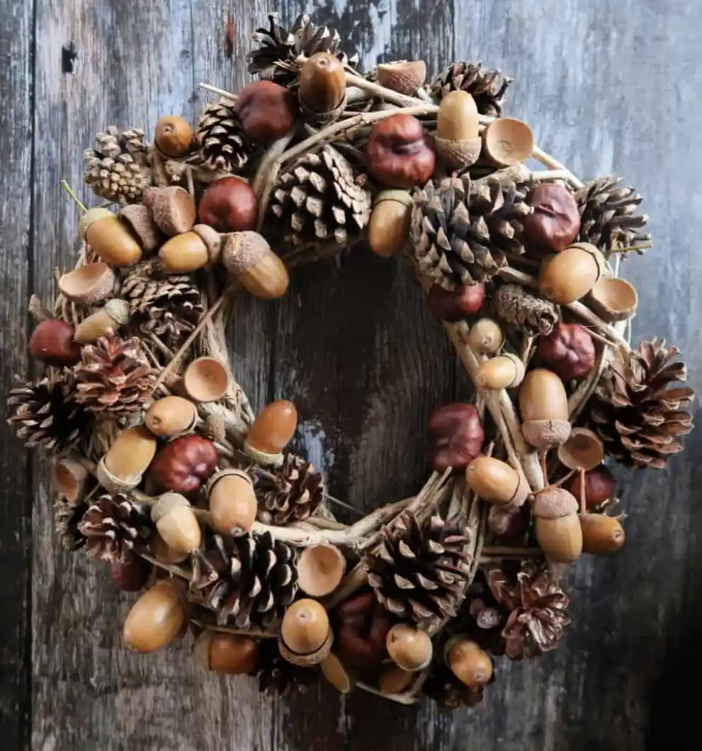A rustic autumn wreath made of twigs, pinecones, acorns, and chestnuts is displayed against a weathered wooden background…