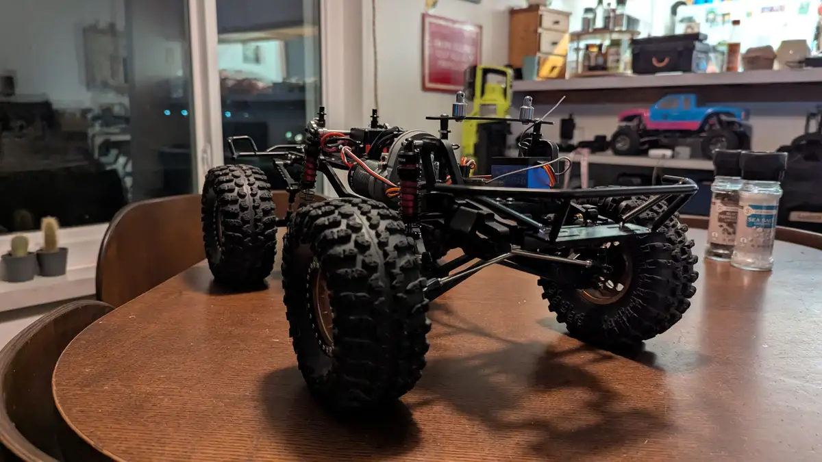 A black remote-controlled monster truck with large knobby tires sits on a wooden table, its exposed chassis and wiring…