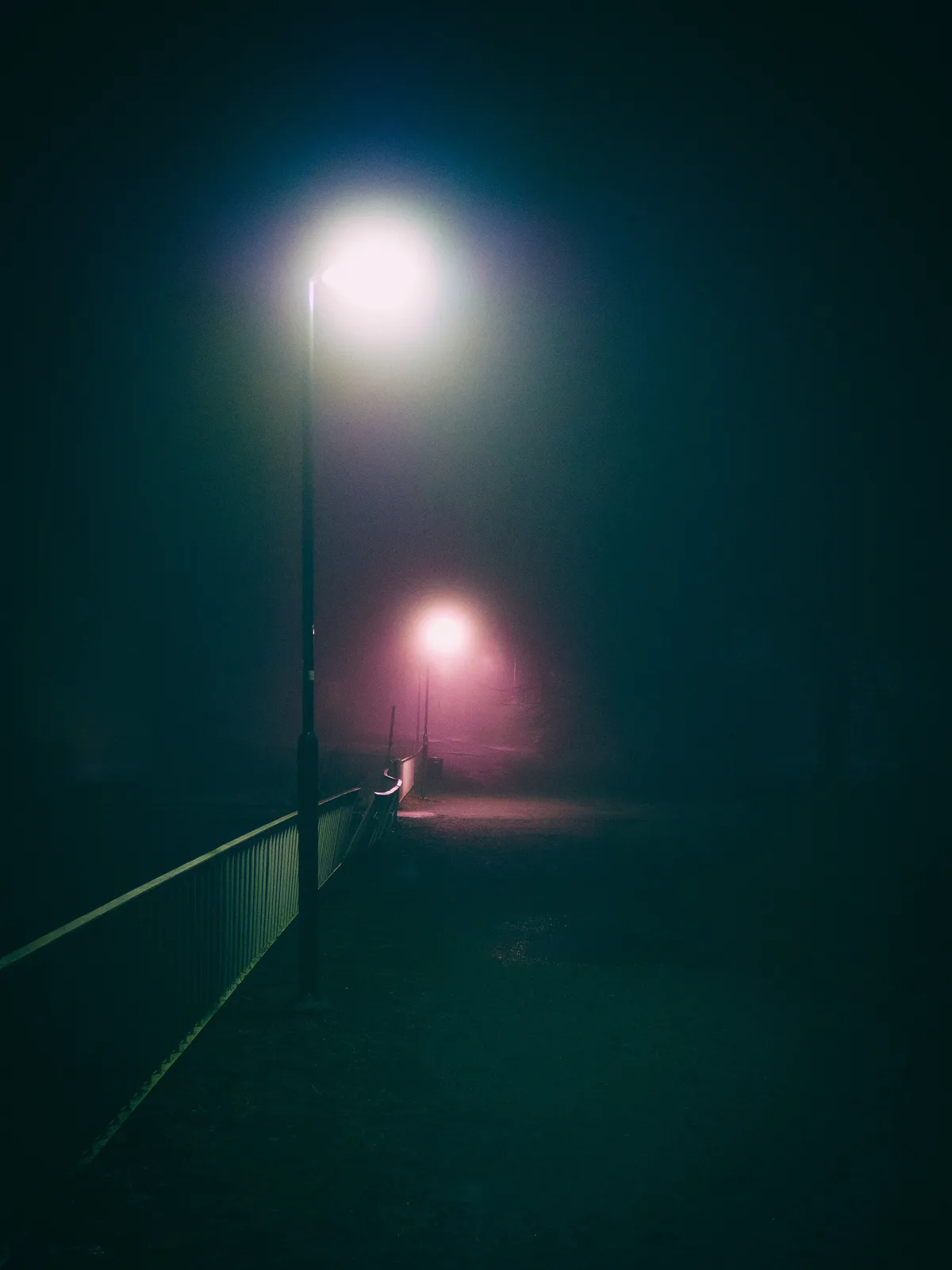 A dark, foggy night scene with two streetlights illuminating a path. The foreground light casts a white glow, while the…