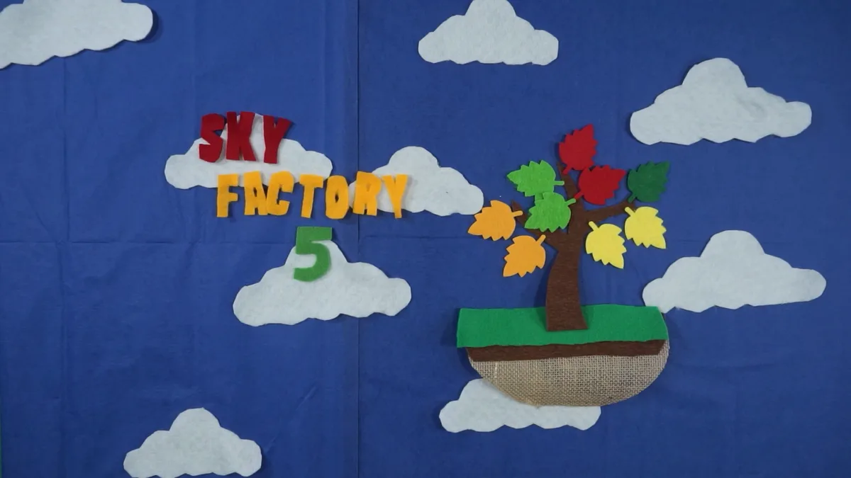 A handmade felt craft displays the words 'SKY FACTORY 5' in colorful letters on clouds against a blue sky background, next…