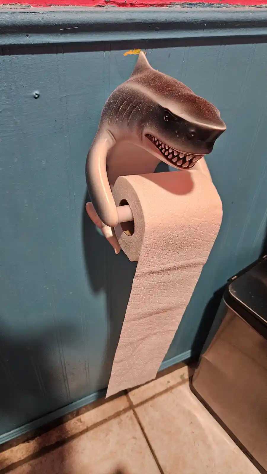 A plastic shark-shaped toilet paper holder with a menacing grin is mounted on a blue wall, holding a roll of toilet paper…