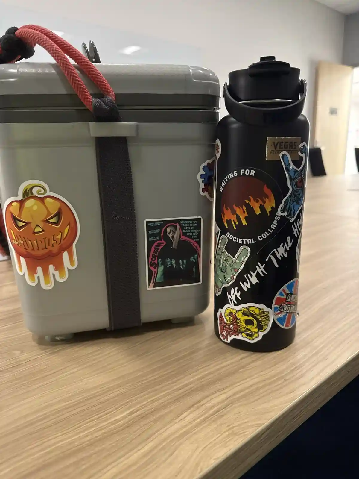 A gray cooler and a black water bottle, both heavily covered in horror, punk, and apocalyptic-themed stickers, sit…
