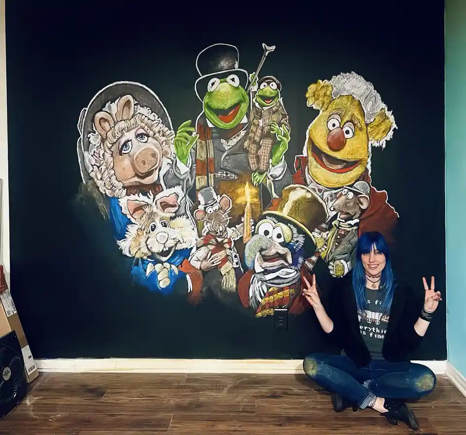 A person with blue hair sits cross-legged in front of a large chalk drawing on a black wall, making peace signs. The artwork…