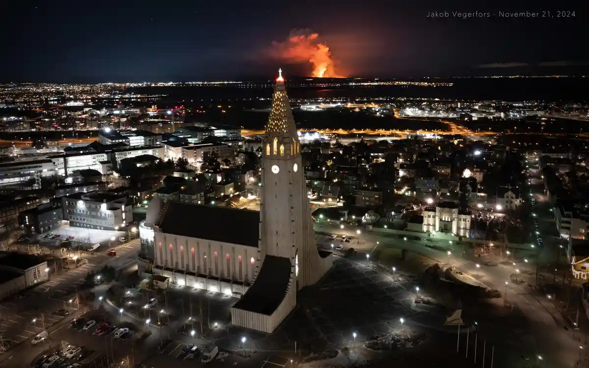 An aerial night photo shows the illuminated Hallgrímskirkja church in Reykjavik, Iceland, with a distant volcanic eruption…