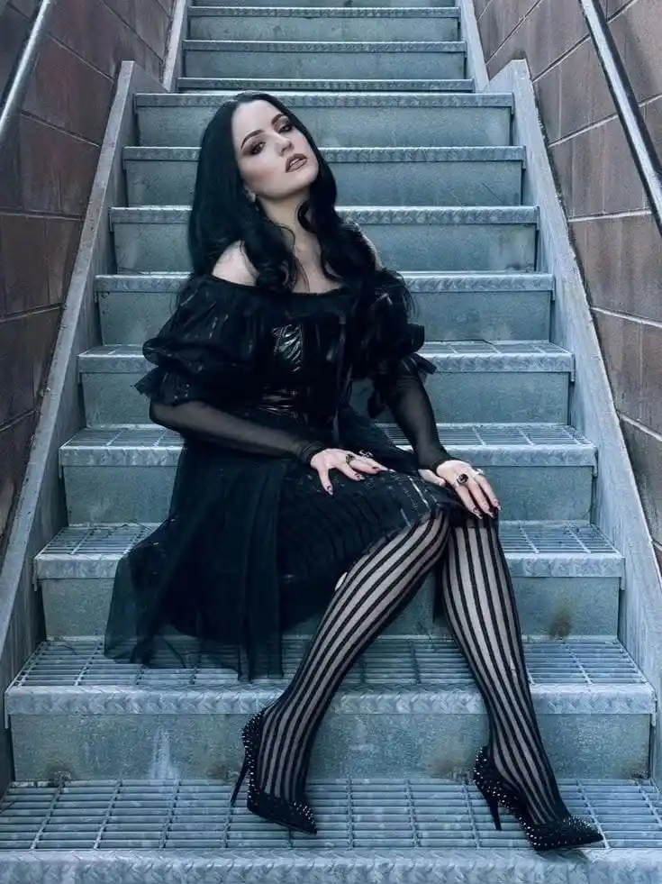 A woman with long dark hair sits on industrial metal stairs wearing a black off-the-shoulder dress, black sheer sleeves…