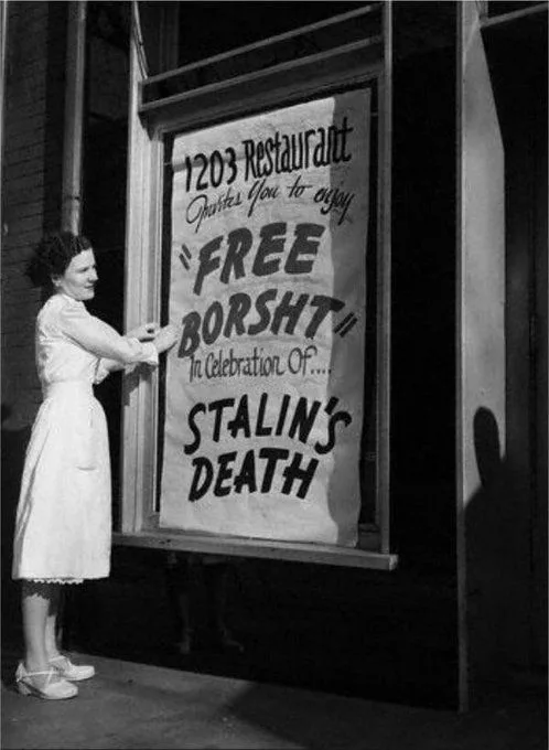 A black-and-white photo shows a woman in a white dress hanging a large sign in a restaurant window. The sign reads '1203…
