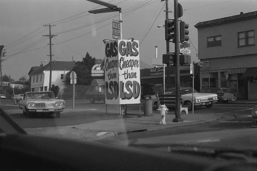 A black-and-white photo of a 1960s street corner with vintage cars and storefronts, featuring a large roadside sign that…