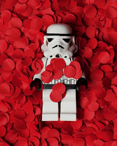 A white LEGO stormtrooper minifigure is surrounded by and covered in red heart-shaped confetti, visually contrasting its…