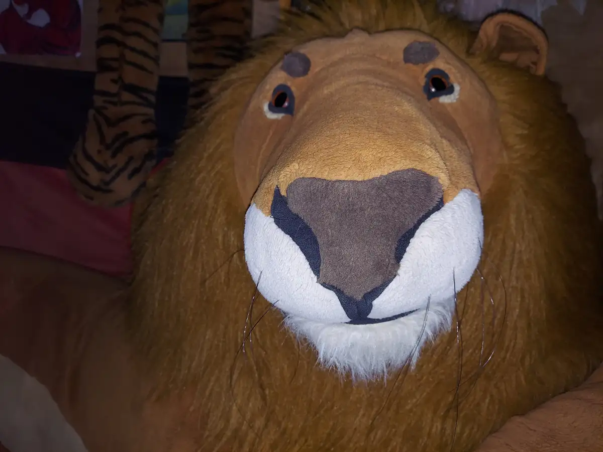 A close-up of a large, brown plush lion toy with a white muzzle and black nose, positioned in a room with a tiger-patterned…