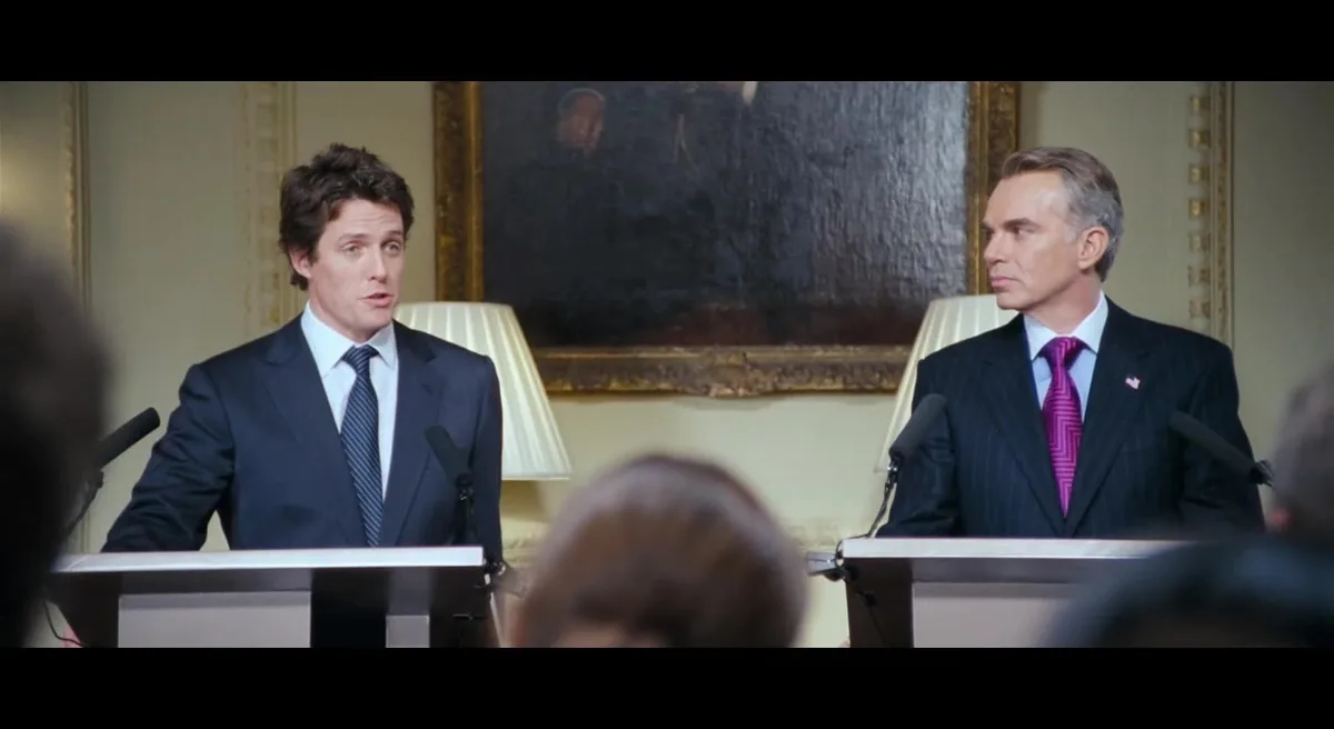 Two men in suits stand at podiums in a formal room, one looking shocked and the other looking sternly disapproving. The man…