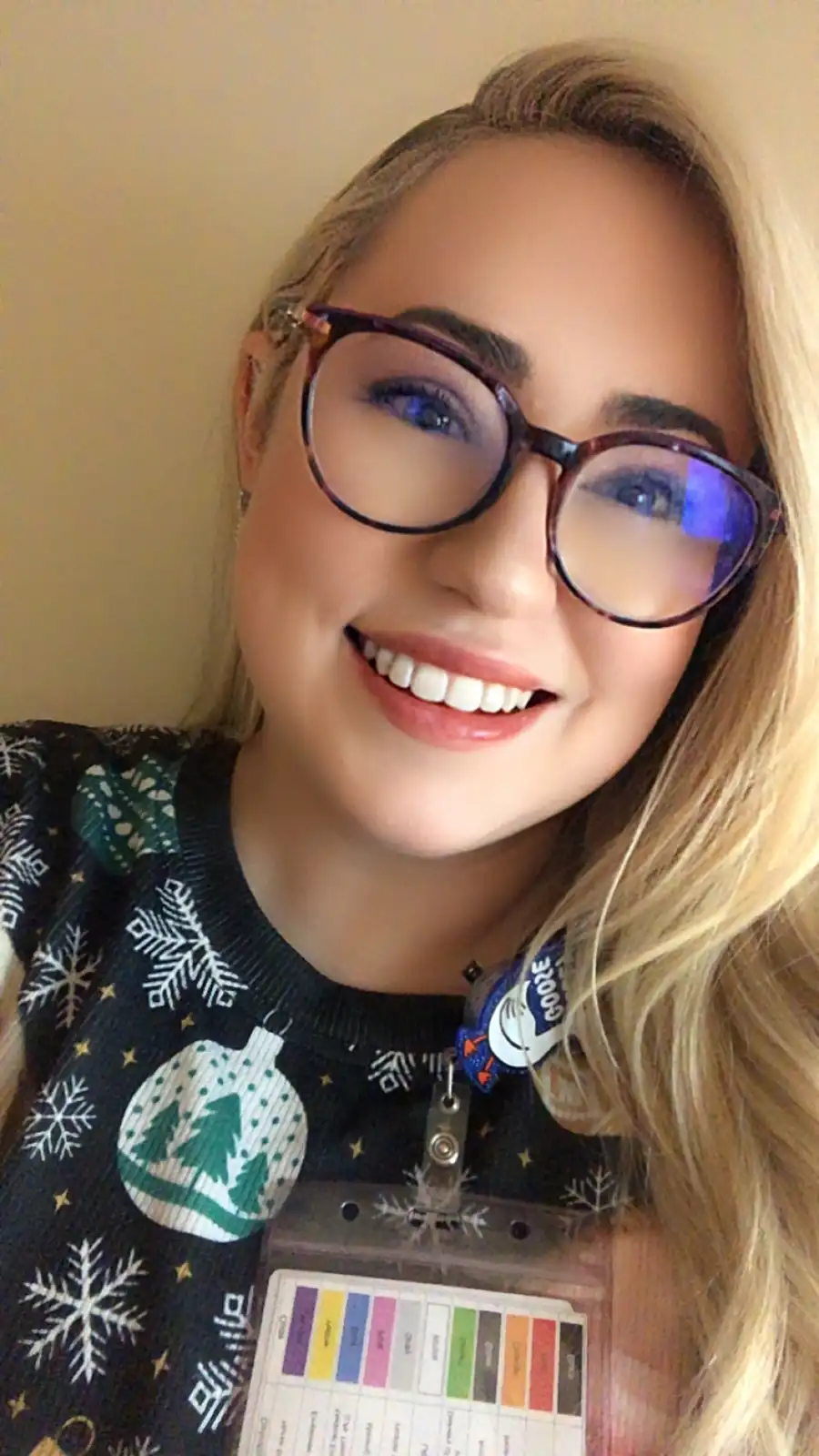 A smiling woman with blonde hair and glasses wears a festive black sweater with white snowflakes and green ornaments. She…