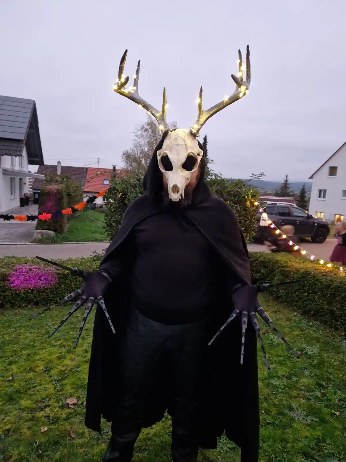 A person in a Halloween costume stands outdoors wearing a black hooded cloak, a deer skull mask with glowing string lights…