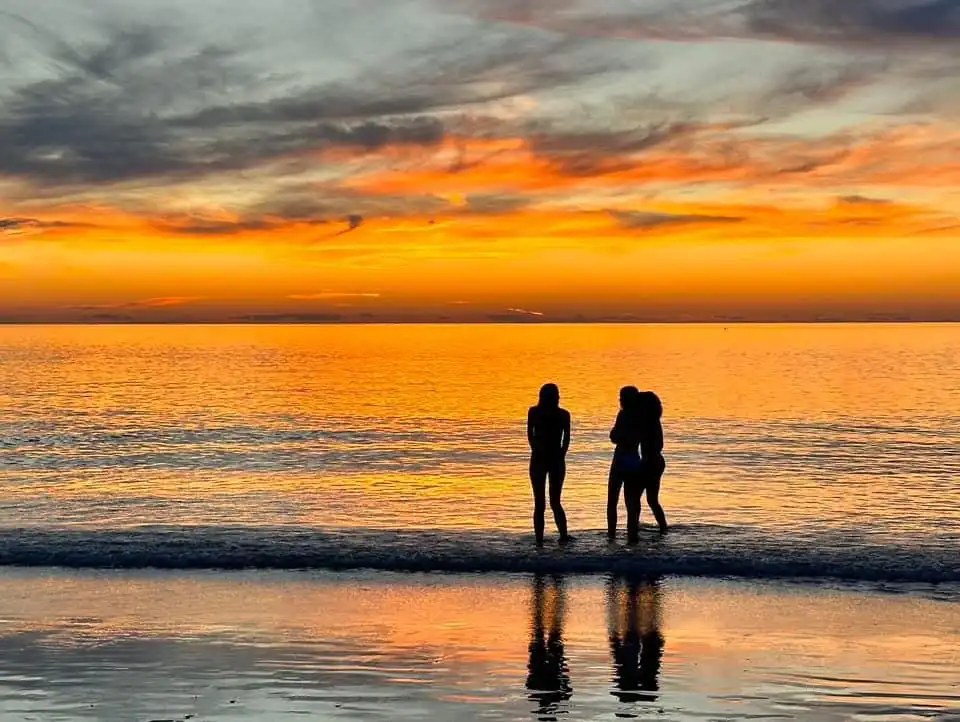 Three silhouetted people stand in shallow ocean water at sunset, with one person hugging another while the third stands…