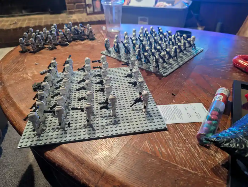 A wooden table holds multiple gray Lego baseplates, each populated with numerous white Lego Stormtrooper figures holding…