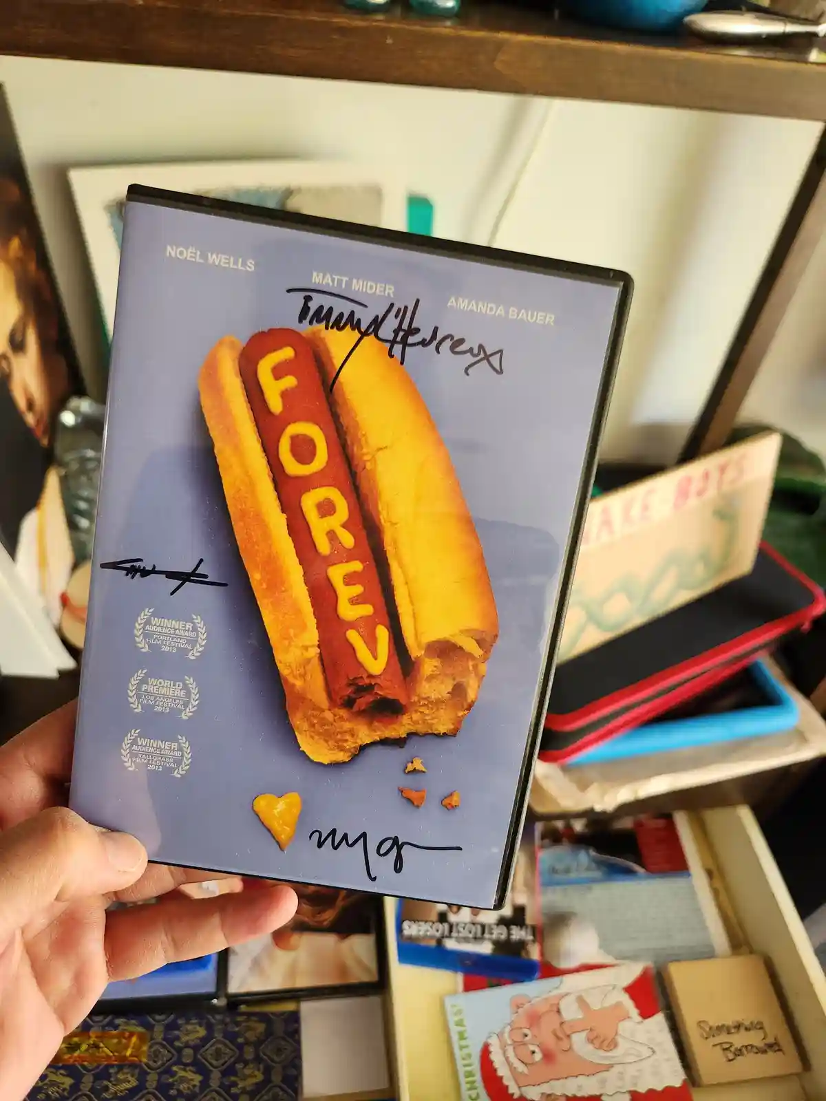 A hand holds a DVD case for the film 'Forever,' featuring a hot dog with mustard spelling 'FOREV' and a heart-shaped mustard…