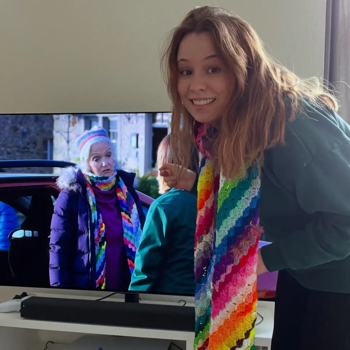 A young woman with brown hair smiles at the camera while wearing a colorful, rainbow-striped crocheted scarf. Behind her, a…