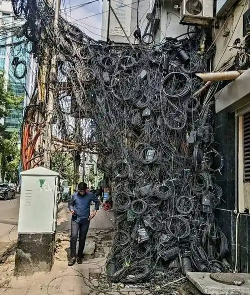 A man stands beside a massive, chaotic tangle of black electrical wires and cables draped over a building facade and utility…