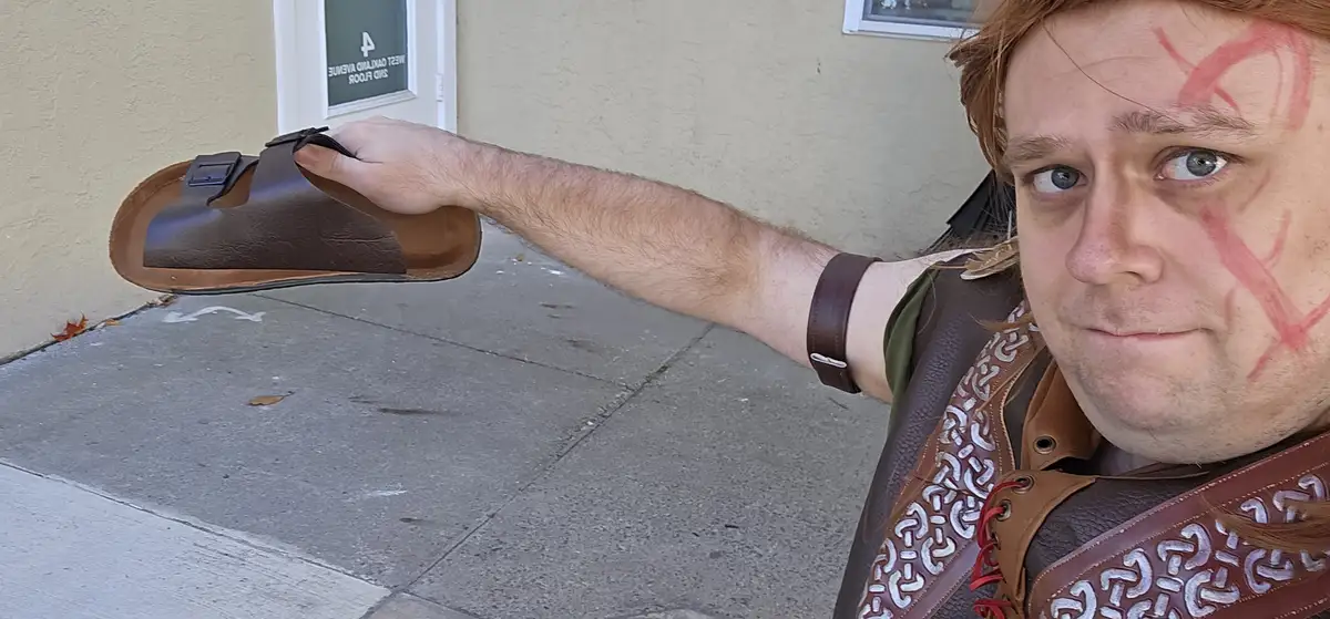 A man dressed in fantasy warrior attire with red face paint holds up a brown Birkenstock sandal as if it were a weapon. He…