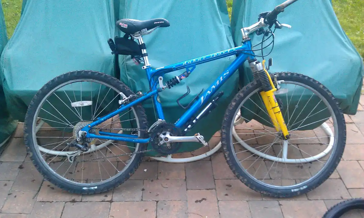 A blue Jamis Dakar Sports mountain bike with a yellow front fork is parked on a brick patio in front of a green tarp-covered…