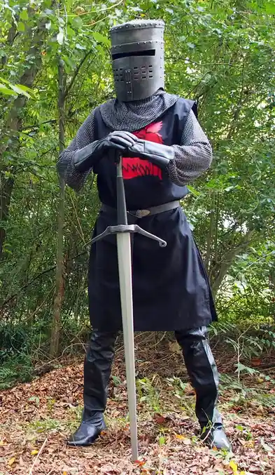 A person in full medieval knight armor, including a helmet with eye slits and chainmail, stands in a forest holding a sword…