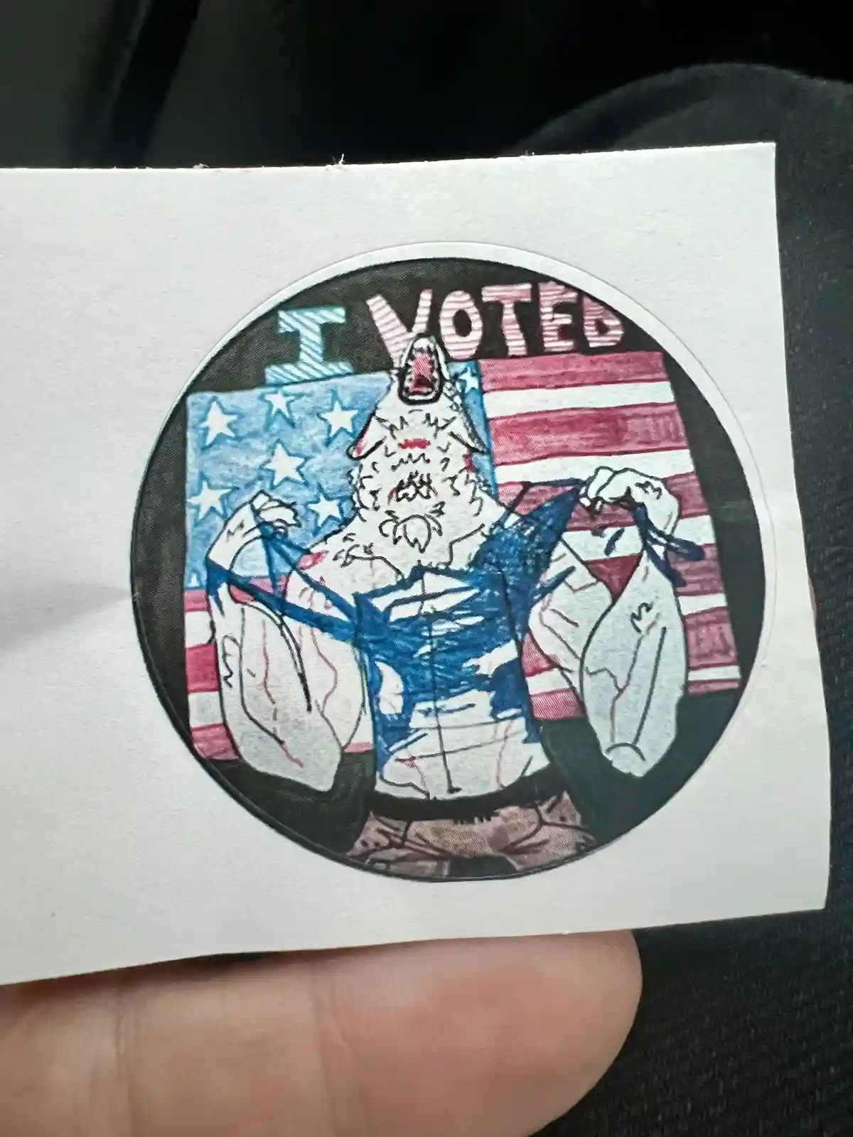 A hand holds a circular sticker depicting a muscular, monstrous, wolf-like creature with a screaming face, flexing its arms…