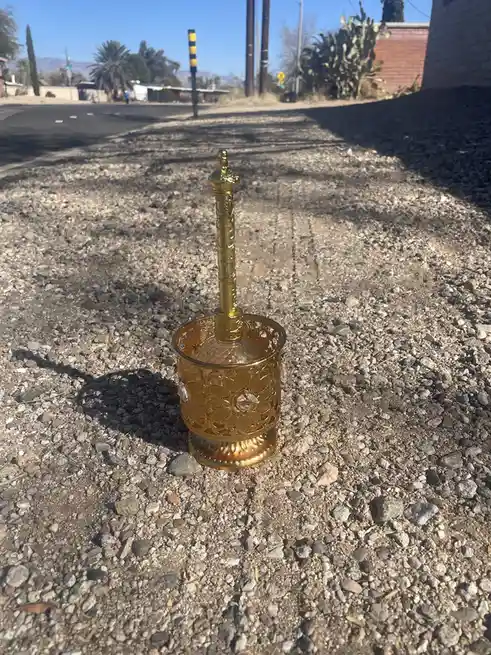 A highly ornate, gold-colored toilet brush with intricate filigree and crystal embellishments sits upright on a gravel…