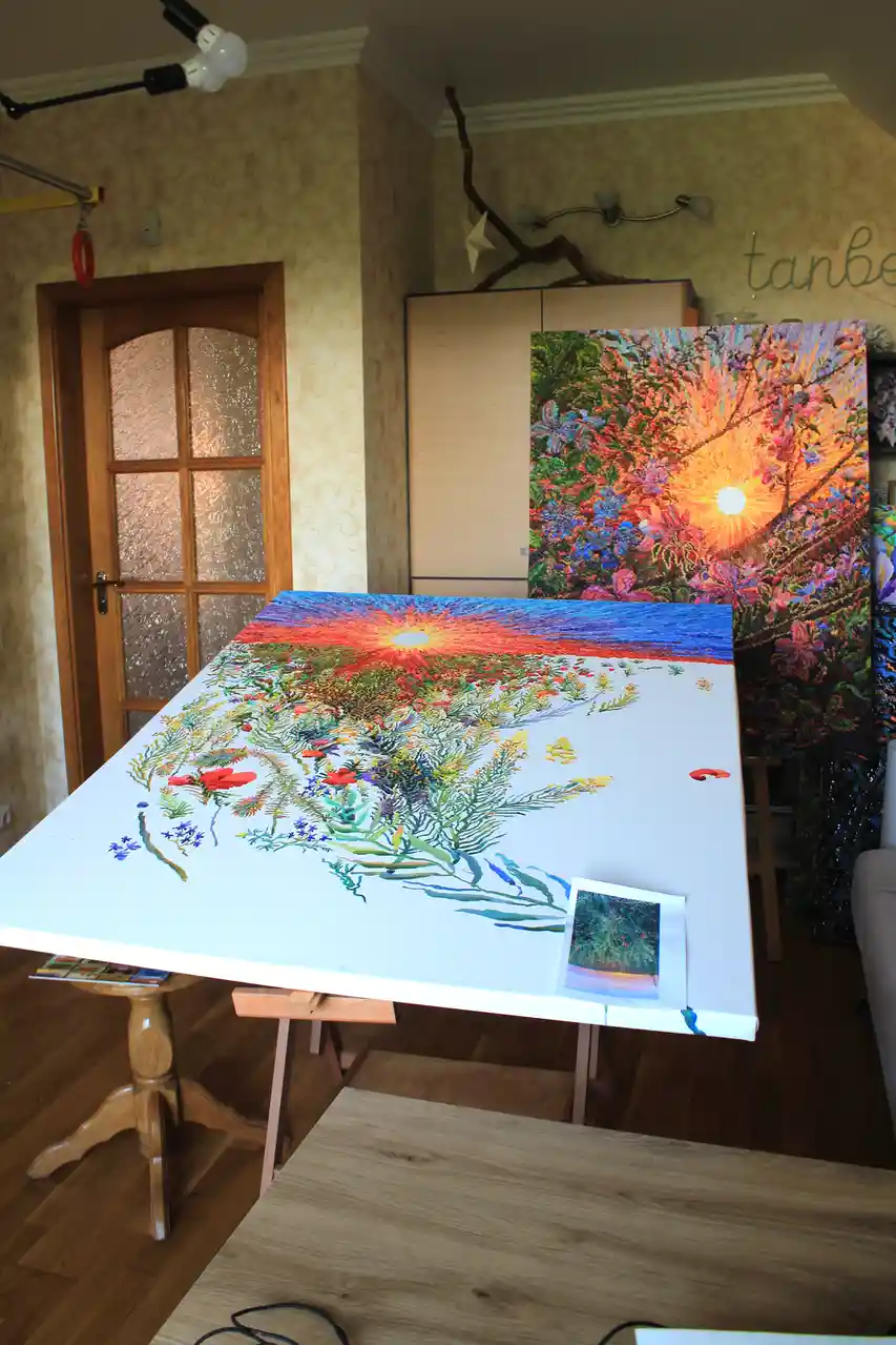 A large canvas on an easel shows a vibrant, partially painted sunset over a field of flowers, with only the lower half…
