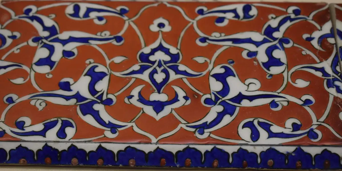 A close-up of an ornate ceramic tile with a repeating pattern of blue and white stylized vines and floral motifs on a…