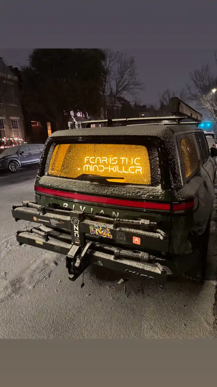 The rear of a dark green Rivian SUV is covered in snow, parked on a snowy street at night. The rear window has a glowing…